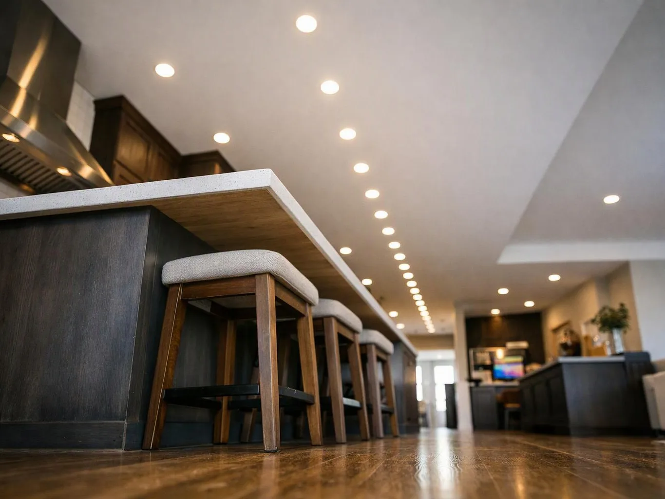 A modern kitchen with a long island, cushioned stools, recessed lights, and a polished wood floor