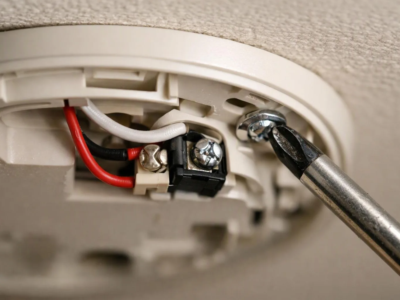 A screwdriver tightens a ceiling smoke detector screw beside red, white, and black wires